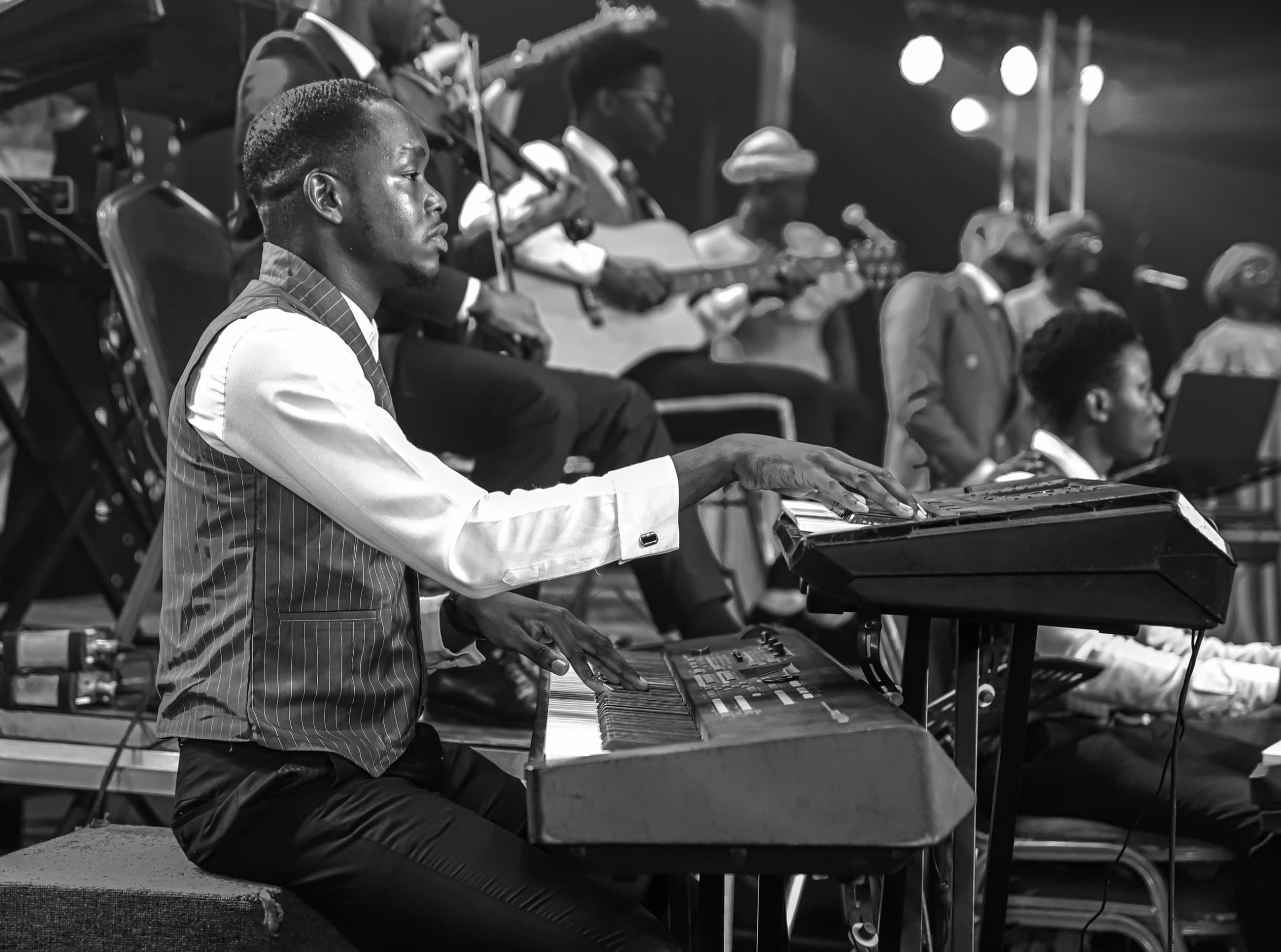 Oladimeji playing two keyboards on stage, with a violinist, guitarist and band behind him, in black and white